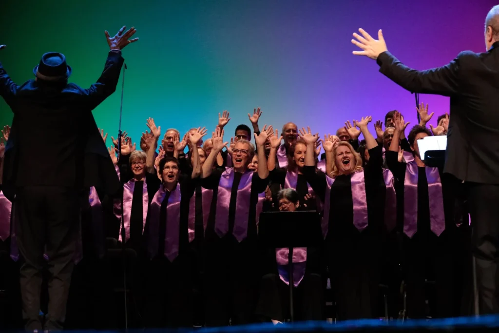 Choir members in purple stoles raising their hands enthusiastically during a performance led by two conductors.