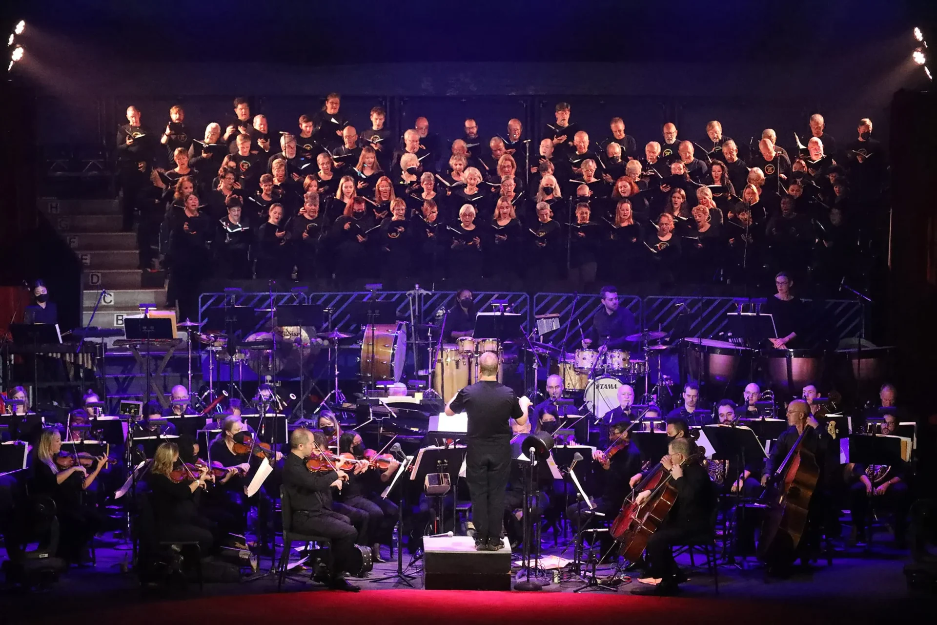 Key Chorale chorus and full orchestra performing together on stage during a Cirque des Voix concert, with percussion, strings, and choir illuminated under dramatic blue and purple lighting.